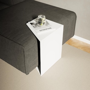 Può includere: Un tavolino bianco dal design minimalista, posizionato accanto a un divano grigio scuro. Un bicchiere d'acqua e un libro con la parola "BAUHAUS" sono sul tavolo. Le linee pulite e i colori neutri del tavolo creano un'estetica contemporanea.