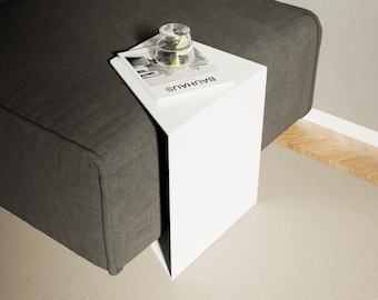 May include: A modern white side table with a minimalist design, placed next to a dark gray sofa. A glass of water and a book with the word "BAUHAUS" are on the table. The table's clean lines and neutral colors create a contemporary aesthetic.
