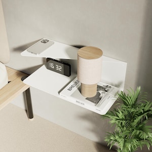 May include: A modern white wall-mounted shelf unit with two triangular shelves. The top shelf holds a smartphone. The bottom shelf displays a digital clock showing 16:32, a lamp, and a book titled "BREUER". A small plant is in the foreground.