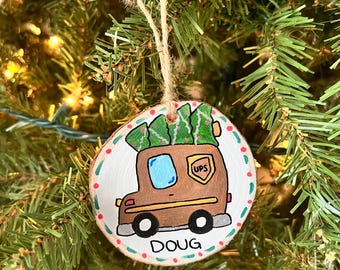 Op de afbeelding: Ronde houten kerstversiering met een bruine UPS-vrachtwagen met groene kerstbomen. De versiering heeft een witte achtergrond met rode en groene stippen langs de rand en de naam "DOUG" eronder. Een touwlus is bevestigd om op te hangen.