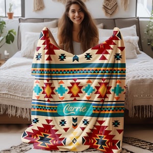 May include: A rectangular throw blanket featuring a cream-colored base with a repeating geometric pattern in red, yellow, teal, and black. The design includes horizontal stripes and diamond motifs. The name Carrie is printed in white script.