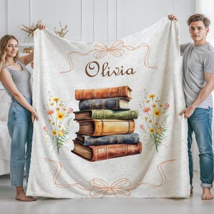 May include: A cream-colored blanket featuring a stack of books with watercolor floral accents and the name "Olivia" in brown script. The blanket has a ribbon design and is held by two people.