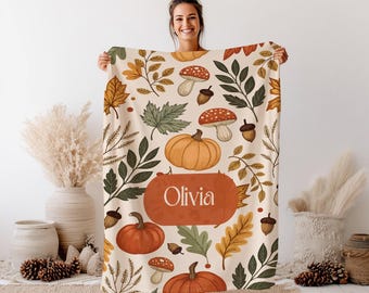 May include: A beige blanket with a fall-themed pattern featuring pumpkins, mushrooms, acorns, and leaves in orange, green, and brown. The blanket has the name "Olivia" in a brown oval.