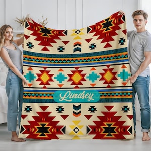 May include: A beige blanket with a Southwestern design featuring red, black, yellow, and turquoise geometric patterns. The blanket has a turquoise banner with the name "Lindsey" in white script. The blanket is held by two people.