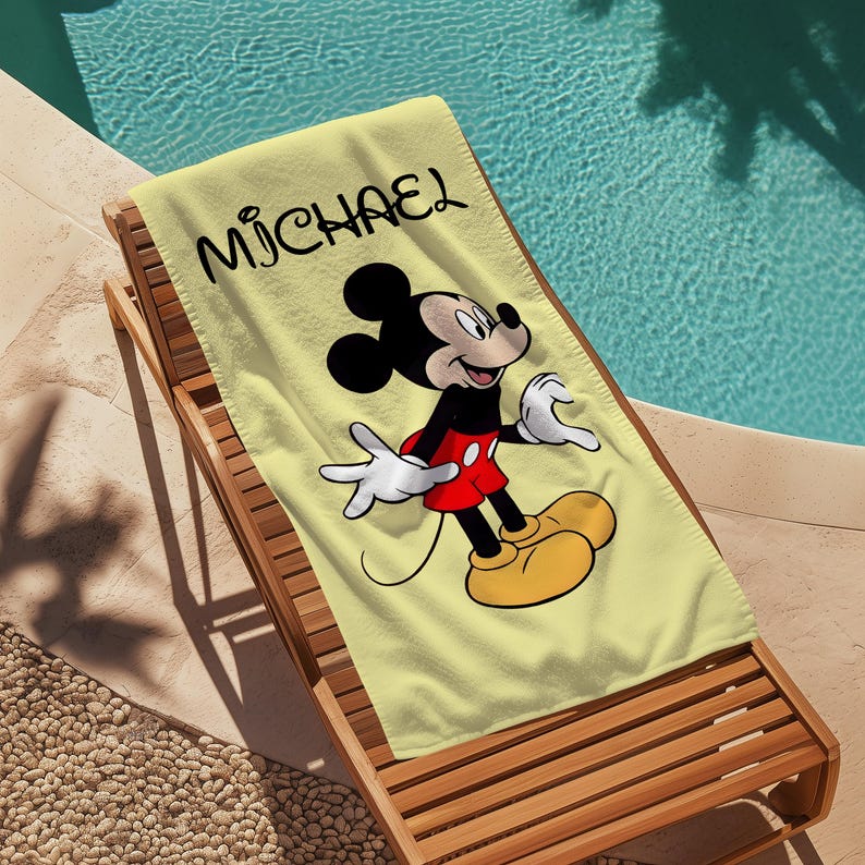 May include: A yellow beach towel featuring a smiling Mickey Mouse cartoon character in a red shorts, black top, and yellow shoes. The name "MICHAEL" is printed in black script across the top of the towel. The towel is on a wooden lounge chair.