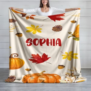 May include: A cream-colored blanket featuring a fall-themed design with pumpkins, leaves, and acorns. The word "SOPHIA" is printed in red. The blanket is a cozy seasonal home decor item.
