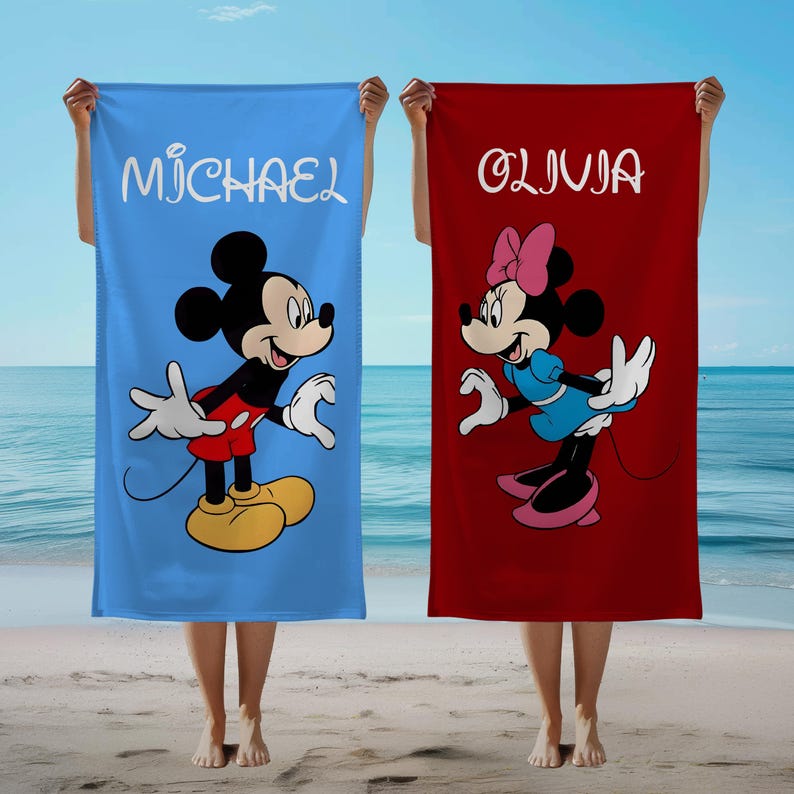 May include: Two beach towels, one blue and one red, are held up against a beach backdrop. The blue towel features Mickey Mouse and the name "MICHAEL" in white script. The red towel displays Minnie Mouse and the name "OLIVIA".