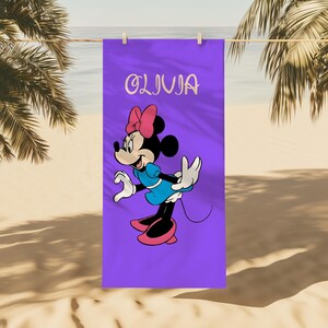 May include: A purple beach towel featuring a cartoon character with a pink bow, blue dress, and pink shoes. The name "OLIVIA" is written in cursive at the top. The towel is hanging on a clothesline with a beach and palm trees in the background.