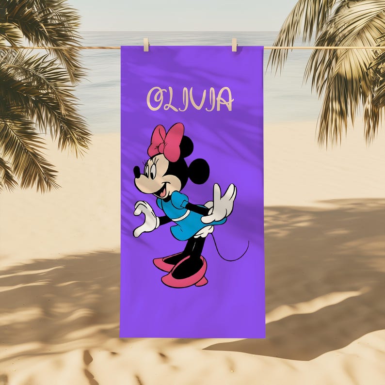 May include: A purple beach towel featuring a cartoon character with a pink bow, blue dress, and pink shoes. The name "OLIVIA" is written in cursive at the top. The towel is hanging on a clothesline with a beach and palm trees in the background.