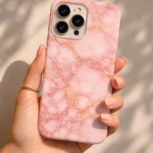 以下が含まれることがあります： ピンクの大理石模様の携帯電話ケース。 金色の模様が入っています。 ケースは長方形で、角が丸く、カメラ用の切り欠きがあります。 手に持たれており、指には金の指輪が見えます。