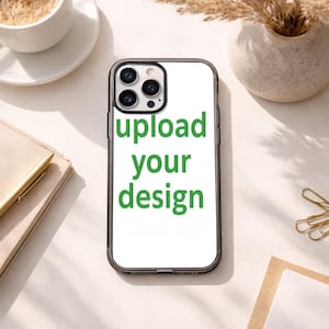 May include: A clear phone case with a white background and the green text "upload your design." The case has a black border and is surrounded by a neutral-toned background with a cup of coffee, a notebook, and decorative items.