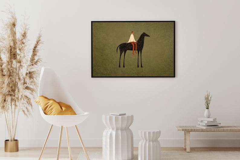 May include: A framed art print depicts a person riding a black horse against a muted green background. The rider wears a white garment and brown pants. The artwork is displayed on a white wall, complemented by a modern chair and decorative elements.