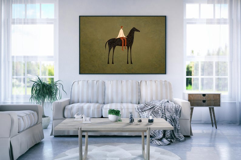 May include: A framed art print depicts a minimalist illustration of a person riding a black horse. The rider wears a white garment and brown pants against a muted olive green background. The artwork is displayed above a sofa in a living room.