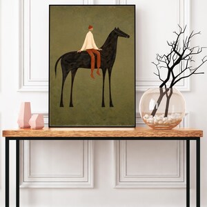 May include: An art print featuring a black horse with a rider in a white shirt, red pants, and a red hat. The artwork is set against a muted green background and framed in black. The print is displayed on a wooden table with a black metal frame, alongside decorative vases.