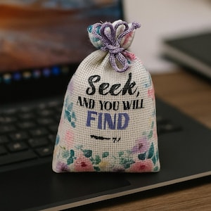 May include: A small, beige fabric bag with a floral pattern and the words "Seek, AND YOU WILL FIND" printed in black and purple. The bag is tied with a purple cord and sits on a laptop.