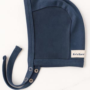 May include: A navy blue baby bonnet with a ribbed texture. The bonnet has a rounded shape with a flap that fastens with snaps. A small label with the text "Ev's Ears" is sewn on the side.
