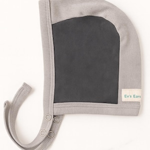 May include: A light gray baby bonnet with a dark gray mesh panel. The bonnet has a ribbed texture and a curved brim. The label on the side reads "Ev's Ears". The bonnet has a chin strap with two snaps.
