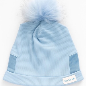 May include: A pale blue ribbed knit beanie hat with a large, fluffy pom-pom. The hat has two light blue patches on the sides and a small white label with the text "Ev's Ears".