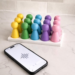 May include: A collection of colorful wooden peg dolls in yellow, green, blue, purple, and pink, arranged on a white tray. A smartphone with a black case displays a sound wave graphic on its screen, resting on a marble surface.