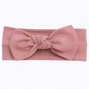 May include: A dusty rose-colored headband with a large bow. The headband is made of a ribbed fabric, with the bow centered on the band. The bow is knotted in the middle, creating a full, textured look. This accessory is suitable for various occasions.