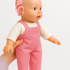 May include: A baby doll with light skin and blue eyes wearing a cream-colored long-sleeved shirt and pink overalls. The doll features a pink hearing aid device worn behind the right ear, which includes a small heart-shaped detail on the ear mold.