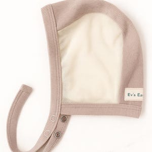 Può includere: Un cappellino da neonato color talpa con pannello frontale in rete e chiusure a pressione. Il cappellino ha una trama a coste e una piccola etichetta con la scritta "Ev's Ear". Design semplice e funzionale per neonati.