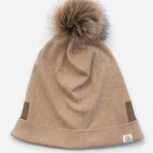 Peut inclure: Un bonnet en tricot côtelé beige avec un gros pompon en fausse fourrure. Le chapeau a un bord replié et de petits patchs marron sur les côtés. Une petite étiquette blanche avec un logo circulaire est cousue sur le côté.