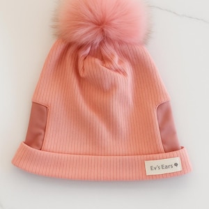 May include: A coral pink beanie with a large, fluffy pom-pom. The hat features a ribbed knit design with smooth side panels and a small tag that reads "Ev's Ears". A stylish and warm winter accessory.