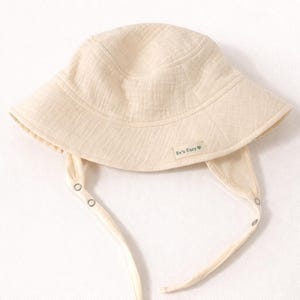 May include: A cream-coloured baby sun hat with a wide brim and chin ties. The hat features a small label that reads "Ev's Ears". The hat is made of a soft, textured fabric and has a simple, classic design.