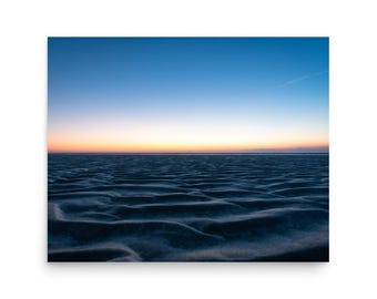 May include: A scenic print of a beach at dusk. The foreground shows rippled sand in shades of blue and gray. The horizon features a gradient of orange and yellow, with a clear blue sky above.