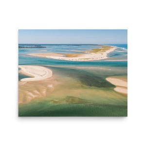 May include: Aerial view of a coastal landscape featuring a long, sandy spit of land surrounded by turquoise and green waters. The sky is a clear blue, and the scene evokes a sense of tranquility and natural beauty. The image is a print.