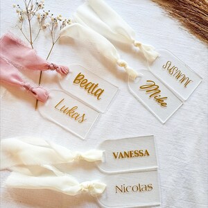 Op de afbeelding: Transparante acryl naamkaartjes met gouden letters en stoffen linten in roze en wit. De kaartjes zijn gepersonaliseerd met namen als Beata, Lukas, Mike, Susanne, Vanessa en Nicolas. Ideaal voor bruiloften of evenementen.
