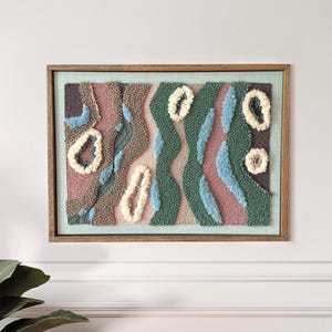 May include: Abstract textile art piece in a wooden frame. The artwork features a textured design with various colors, including pink, green, blue, and brown. The design includes organic shapes and patterns, creating a unique wall hanging.
