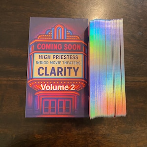 May include: A deck of cards with iridescent edges and a box featuring a vintage movie theater design. The box reads "COMING SOON," "HIGH PRIESTESS," "INDIGO MOVIE THEATERS," and "CLARITY Volume 2." The color scheme includes blue, red, and gold.