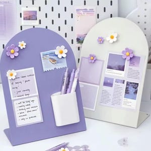 Könnte beinhalten: Zwei dekorative Schreibtisch-Organizer in Lavendel und Weiß, jeweils mit gewölbter Oberseite und Sockel. Der lila Organizer hat einen weißen Stifthalter und hält Notizen und Stifte. Beide haben magnetische Gänseblümchen und zeigen Fotos.