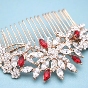 Könnte beinhalten: Ein goldfarbener Haarkamm mit einem dekorativen Blumenarrangement, das mit klaren weißen Kristallen und marquise-geschliffenen roten Steinen besetzt ist. Der Metallkamm hat mehrere Zinken und ein symmetrisches Muster aus Edelsteinen.