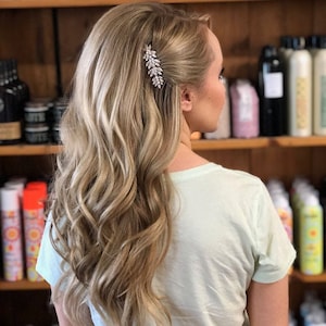May include: A woman with long, wavy, light blonde hair styled with a sparkly hair clip. The hair is parted on the side and pulled back slightly on one side.