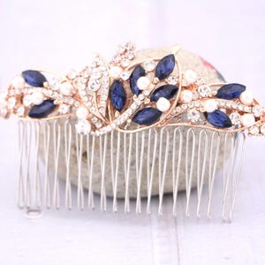 May include: A hair comb featuring a gold-tone metal base adorned with marquise-cut dark blue crystals, round white faux pearls, and small clear rhinestones arranged in a decorative floral and leaf-inspired pattern across the top edge.