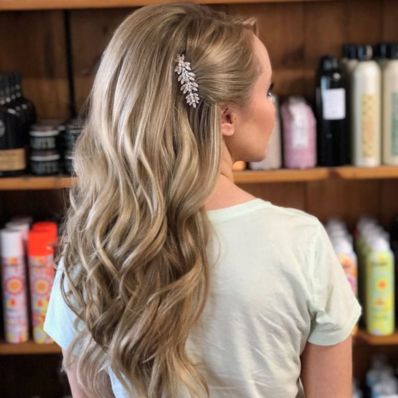 May include: A woman with long, wavy, light blonde hair styled with a sparkly hair clip. The hair is parted on the side and pulled back slightly on one side.