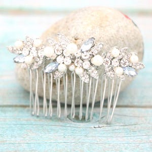 May include: Three silver hair combs with floral designs. Each comb features clear and light blue crystals, and white faux pearls. The combs are resting on a light gray stone, against a light blue wooden surface.