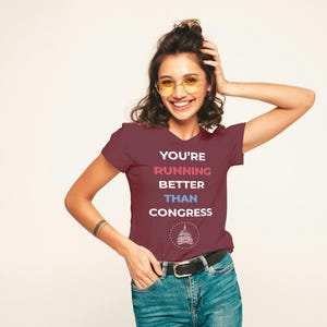 Pu&ograve; includere: T-shirt bordeaux con la scritta "YOU'RE RUNNING BETTER THAN CONGRESS" in bianco, rosso e blu. La maglietta presenta una grafica di un edificio. La persona indossa jeans blu e una cintura nera.