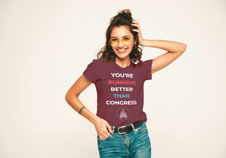 Pu&ograve; includere: T-shirt bordeaux con la scritta "YOU'RE RUNNING BETTER THAN CONGRESS" in bianco, rosso e blu. La maglietta presenta una grafica di un edificio. La persona indossa jeans blu e una cintura nera.