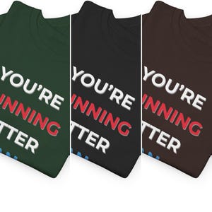 Pu&ograve; includere: Tre magliette in diversi colori: verde scuro, nero e marrone. Ogni maglietta mostra il testo "YOU'RE WINNING BETTER" in lettere bianche e rosse. Le magliette sono piegate, mostrando il testo.