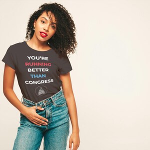 Pu&ograve; includere: T-shirt grigio scuro con la scritta "YOU'RE RUNNING BETTER THAN CONGRESS" in rosso, blu e bianco. La maglietta presenta una grafica di un edificio. La persona indossa jeans blu.
