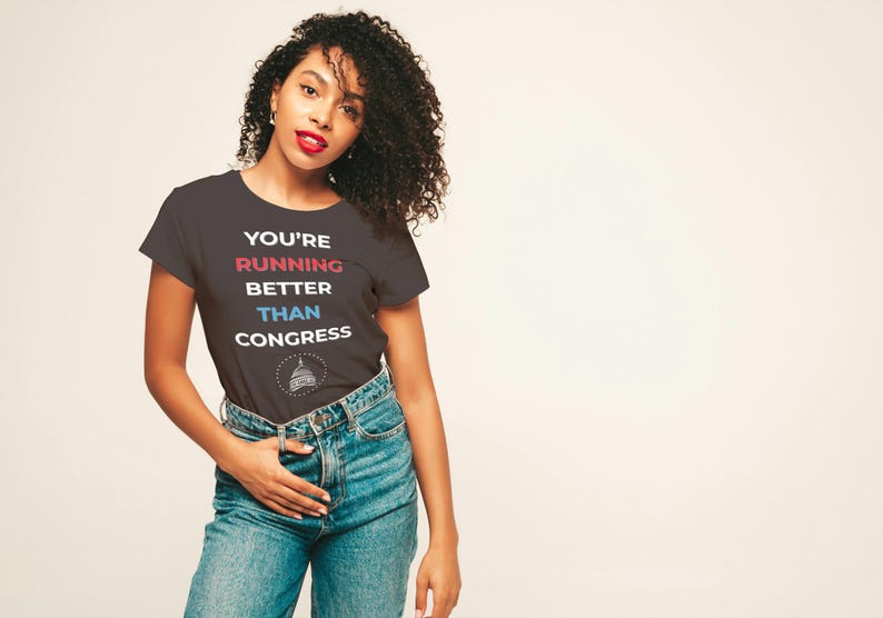Pu&ograve; includere: T-shirt grigio scuro con la scritta "YOU'RE RUNNING BETTER THAN CONGRESS" in rosso, blu e bianco. La maglietta presenta una grafica di un edificio. La persona indossa jeans blu.