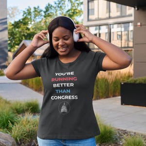 Pu&ograve; includere: T-shirt nera con la scritta "YOU'RE RUNNING BETTER THAN CONGRESS" in rosso, bianco e blu. La maglietta presenta una grafica del Campidoglio degli Stati Uniti. La persona indossa jeans blu e cuffie bianche.