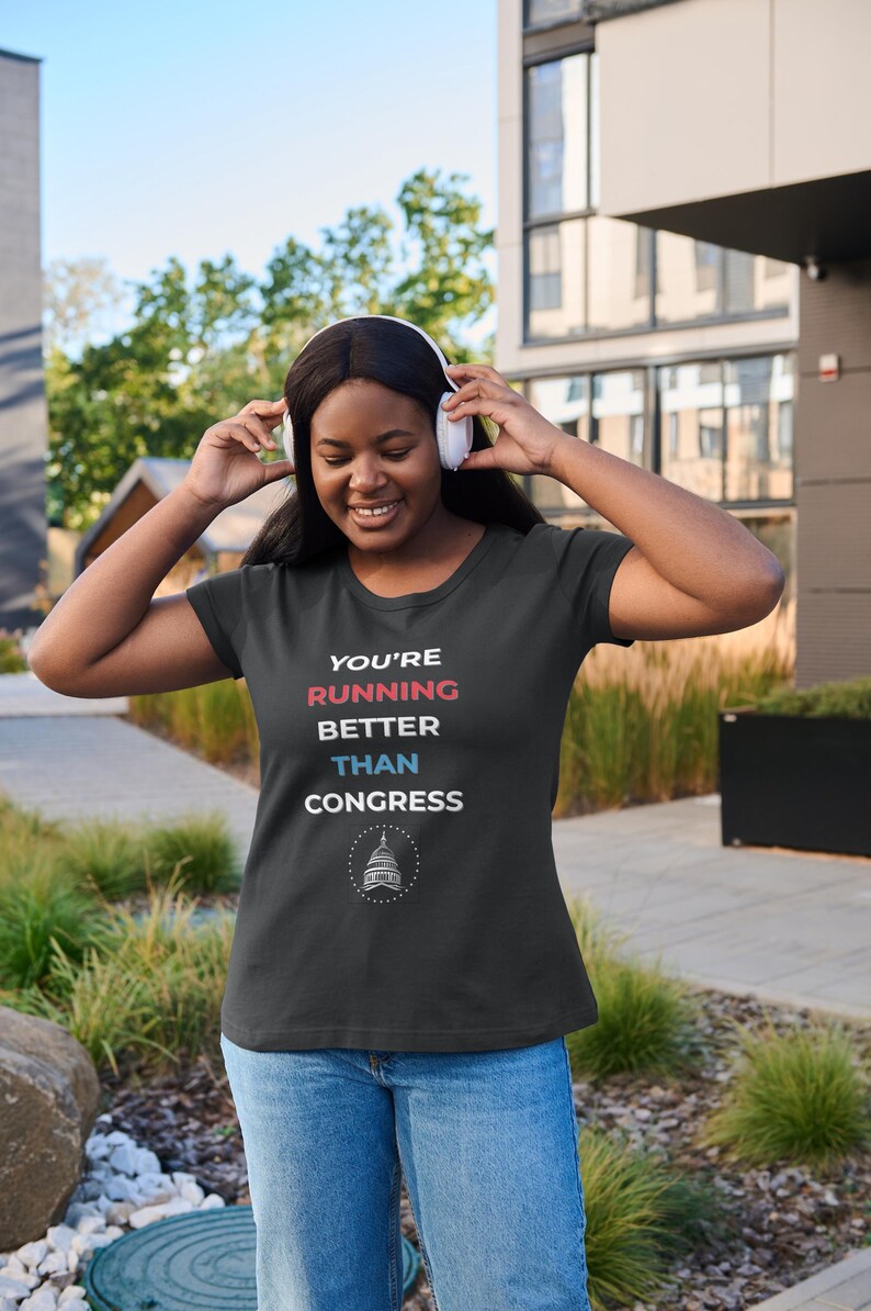 Pu&ograve; includere: T-shirt nera con la scritta "YOU'RE RUNNING BETTER THAN CONGRESS" in rosso, bianco e blu. La maglietta presenta una grafica del Campidoglio degli Stati Uniti. La persona indossa jeans blu e cuffie bianche.