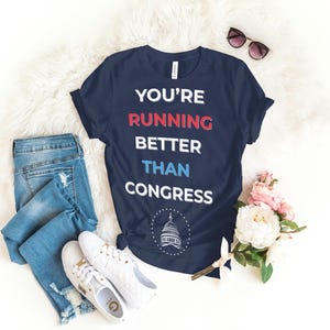 Pu&ograve; includere: T-shirt blu navy con la scritta "YOU'RE RUNNING BETTER THAN CONGRESS" in bianco, rosso e blu. L'immagine include jeans blu, scarpe da ginnastica bianche e occhiali da sole. &Egrave; presente anche un bouquet di fiori rosa e bianchi.