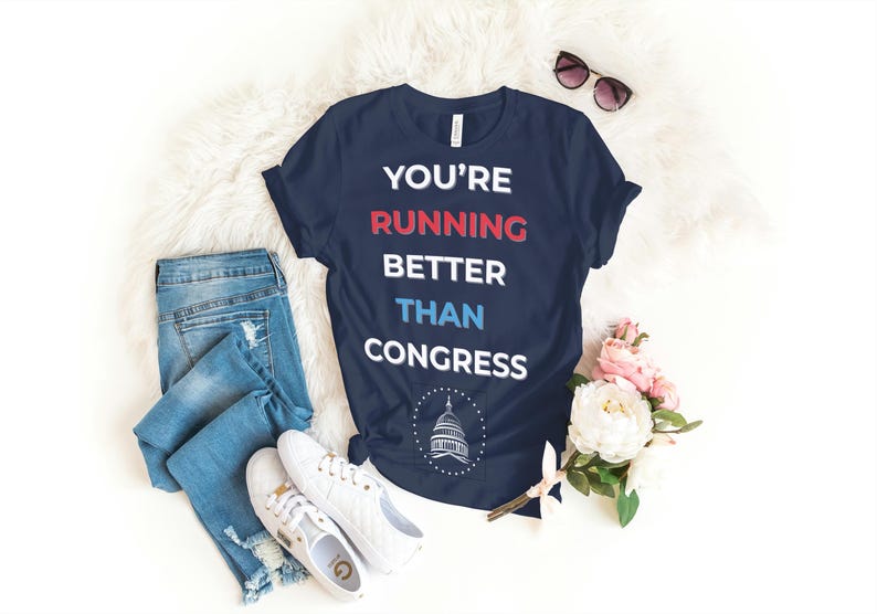 Pu&ograve; includere: T-shirt blu navy con la scritta "YOU'RE RUNNING BETTER THAN CONGRESS" in bianco, rosso e blu. L'immagine include jeans blu, scarpe da ginnastica bianche e occhiali da sole. &Egrave; presente anche un bouquet di fiori rosa e bianchi.