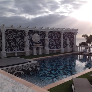 May include: A luxurious outdoor swimming pool with a decorative mosaic tile design. The pool area features white columns, statues, and a patterned wall. The sky is cloudy with a glimpse of the sun.